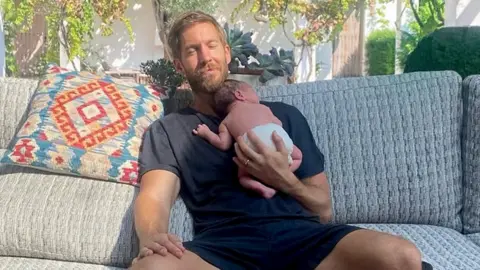 Calvin Harris/Instagram A man sits back on an outdoor sofa, holding a newborn baby against him, supported with one hand as the sun shines on him. The man's eyes are closed as he smiles, tenderly.