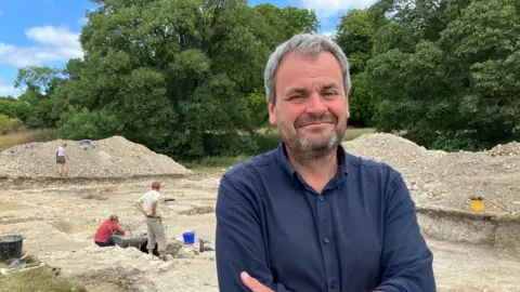 BBC Dr Hugh Willmott smiling for the camera beside the excavated Great Hall at Cerne Abbey. He has dark grey short hair and a full dark beard and is wearing a Navy blue open-collar shirt. Behind him is the a large chalky excavation area where two people are working.