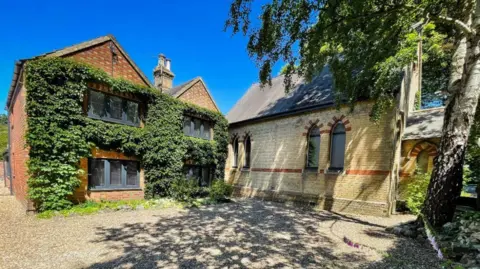 Andy Dransfield A two-storey brick building covered in thick green ivy next to a lighter-coloured brick building with arched windows. The buildings are separated by a gravel driveway. The sky is blue and the trees are casting shadows on the ground.