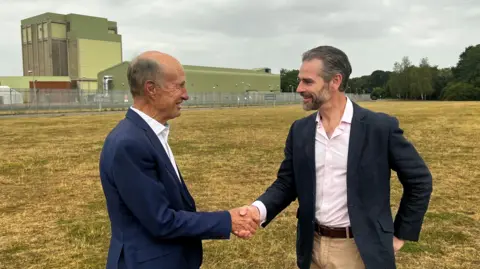 BBC Two men shake hands, standing on a large empty field.  Both wear blue blazers, the one on the right wears a pink shirt and has a grey beard. Behind them is an old green and brown concrete building, the former nuclear reactor.