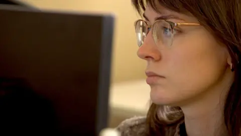Young woman wearing glasses looks at. computer monitor screen.