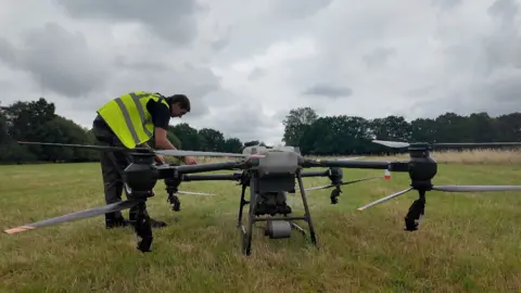 BBC A man wearing a yellow fluorescent safety jacket leans over a large drone to fold out a rotor arm