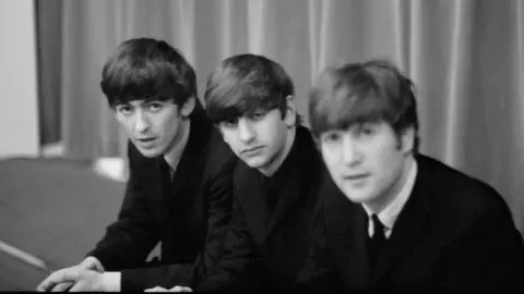 Paul McCartney, Courtesy Gagosian Titled: George, Ringo, and John backstage at The Beatles Christmas Show, Finsbury Park Astoria, London, December 1963. The three bandmates are sat down and look down the lens of the camera. They wear dark suits and are captured in black and white film.