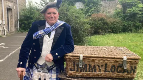BBC A man in a flamboyant, bright tartan dress outfit is leaning on a stick and against a large basket with Jimmy Logan written on the front. He is in a garden and he looks happy.