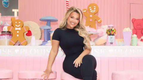 P.Louise Paige has long blonde hair which has been wanded into curls. She is wearing an all black gym set and is sat on a pink bar stool in front of an all pink bar. The bar has the P.Louise branding across it and a number of decorative items including a gumball machine and gingerbread men.