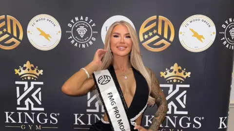 Supplied Victoria Emily, wearing a black dress and standing in front of a backdrop featuring a logo for Kings Gym. She is looking into the camera and smiling.