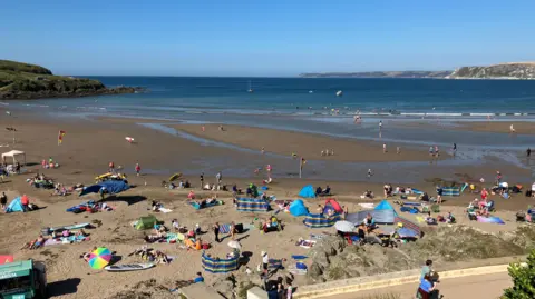 A view over the beach at Bigbury-on-Sea. There are beach umbrellas and windbreaks on the sand. The sky is a clear blue with no cloud. 