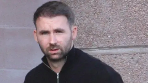 Tim Bugler James Keatings outside Falkirk Sheriff Court. He is only visible from the shoulders up. He is wearing a black quarter zip jumper which is zipped most of the way up. He has short, dark hair and a short, dark beard.