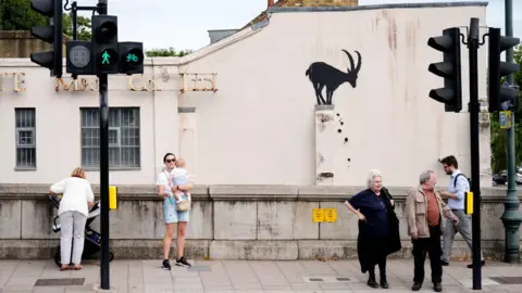 PA Media People walk past a depiction of a defecating goat which has been spray painted on the wall of a house in London.