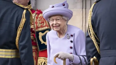 PA Media The late Queen Elizabeth wearing a lilac suit and hat , white gloves and holding a walking stick. She is wearing pearls and a broach. She is flanked by three men in royal livery but their faces are not visible.  