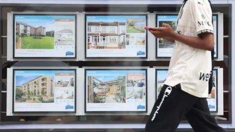 NEIL HALL/EPA/Shutterstock A man walks past an estate agent window filled with six bright digital screens displaying homes.