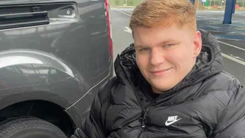 Laura Cooper Ralph Pearce has short ginger hair and is wearing a black coat while sitting in a wheelchair in a car park. He is smiling.