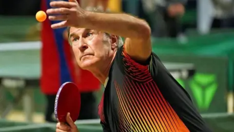 Tim Diss John Poysden staring at an orange table tennis ball he has just thrown in the air. He is holding a red paddle and a black and red top.