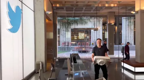 AFP via Getty Images Elon Musk carries a sink into the reception of the headquarters of X, then known as Twitter, on the wall is the old Twitter logo.