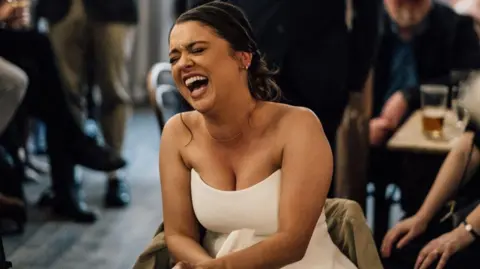 Joseph Dewey A woman wearing a white wedding dress is laughing while sat on a chair