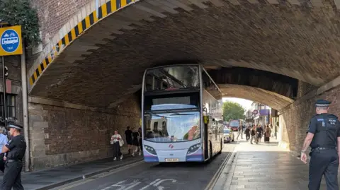 Tracey Franklin A blue double-decker bus going under a low tunnel, a police officer is on the path to the right