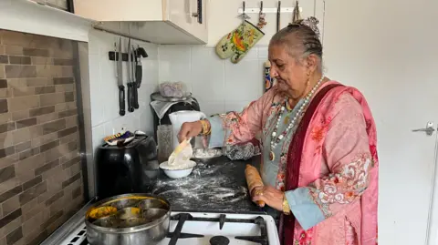 Krishna Singh wearing a blue and pink floral patterned sari, styled with a long white beaded necklace, gold earrings, and thick gold bangles. She is standing in her kitchen, using a wooden rolling pin to roll out dough on the black floured countertops. Her kitchen has brown tiles over the stove, and a magnetic knife holder above the toaster.