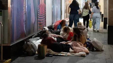 Facundo Arrizabalaga/MyLondon People lying down on blankets and pillows in front of a shop with fashion adverts in the building's windows. with large glass windows. A group walk past in the background.