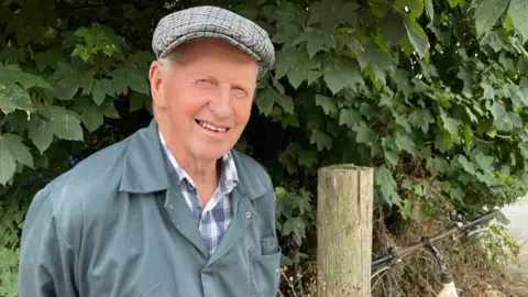 An older man in green overalls smiling at the camera. He is standing in front of a green bush.