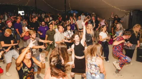 Ania Shrimpton A group of people dancing underneath a marquee. It is dark outside and there are strings of lights draped across the top of the marquee.