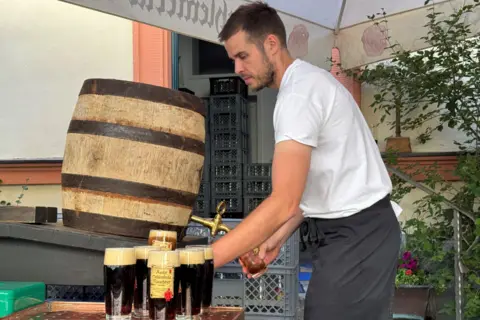 BBC A barman under a bar umbrella pours a dark beer from a barrel. He is wearing a white t-shirt and an apron and is photographed in profile. There are about six beers on the table beside him.