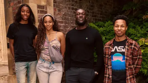Louis Browne The rapper Stormzy with student Destiny, Drew and Dimario alongside him. They stand outside a brick building with shrubbery.