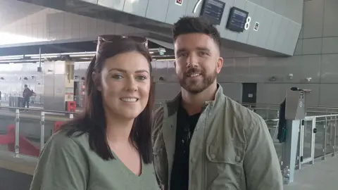 A man and woman smile to the camera. Behind them is a train platform. The woman who stands on the left is wearing a green t-shirt, she has long brown hair with sunglasses resting on the top of her head. The man is wearing a green jacket with a black t-shirt under. He has short brown hair and a short brown beard. 