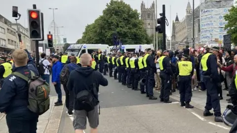 BBC "A street protest scene with a line of police officers in high-visibility vests forming a barrier. Police vans are parked in the background, and a historic building under renovation is visible on the right, with scaffolding and construction materials present.