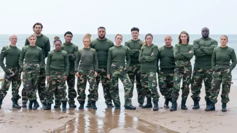 Pete Dadds | Channel 4 The celebrity recruits, wearing military fatigues, line up on a beach with their backs to the sea. They are stood in two rows, some with their arms crossed, with serious expressions. 