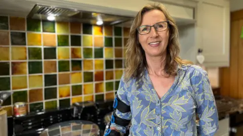 Jayne Jones has brown/blonde hair and is wearing a blue shirt. She is standing in a kitchen.