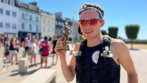 James stands in a sunny urban street holding a small trophy in their left hand. They wear a black athletic vest with two water bottles on the front and a circular emblem patch. Behind them, people walk along the street lined with buildings under a clear blue sky