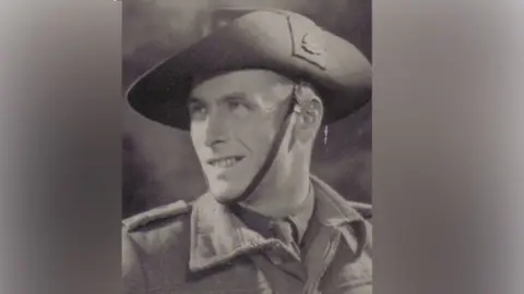 Harkness family A black and white photo of Jack whose head is turned to the side, looking up slightly with a smile. He wears a wide brimmed hat with a strap under his chin, and a thick army jacket over the top of a shirt and tie.