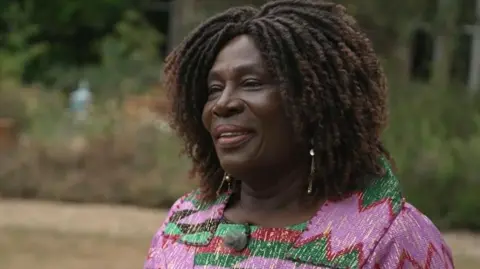 BBC One Show Abigail Akweley smiles and stands in a garden at Cambridge University. She wears a bright pink and green jacket.
