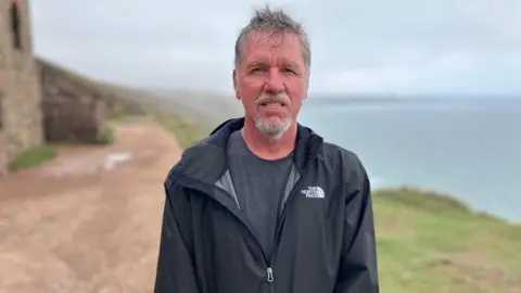 BBC Richard Johnston smiling at the camera and wearing a black coat with a grey t-shirt. He is stood on a coastal path which has a sea view.
