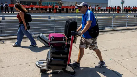 Reuters Man in blue short-sleeve shirt and camo shorts pushes trolley stacked with suitcases next to a woman with long hair, jeans, red tank top and backpack who pulls a gray suitcase