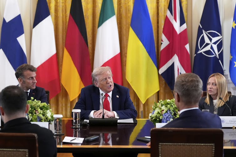 Several people seated around a table listening to a hulking rotund man with orange-ish grey hair. A row of flags is behind him.