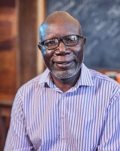 A bald-headed African man in glasses and a striped, collared shirt smiles at the camera.