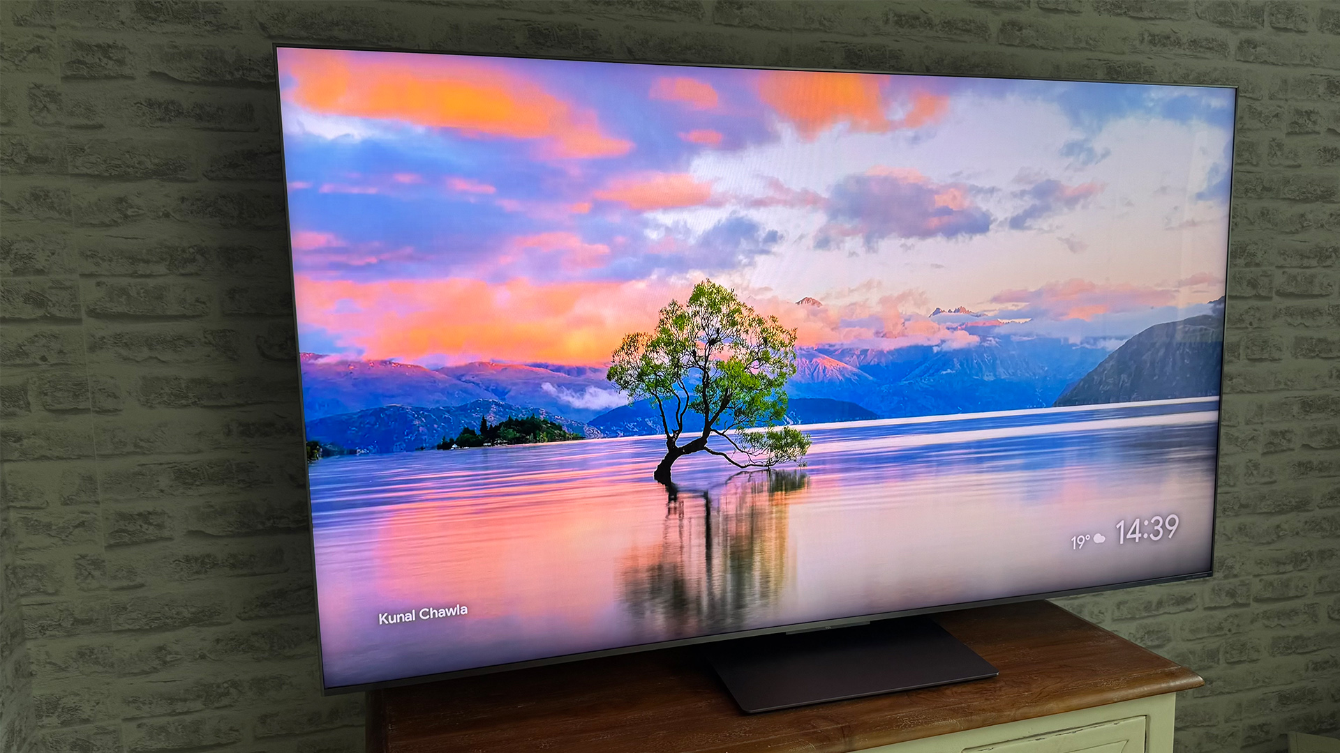 TCL 65C8K LCD TV on wooden cabinet in front of brick wall, on screen is lake and tree