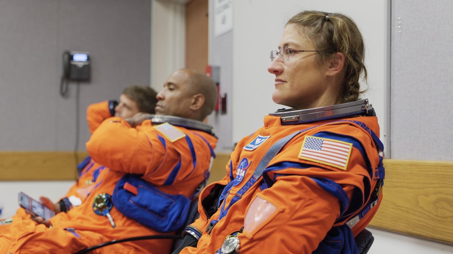 three people in orange flight suits sit inside a drab-looking office room