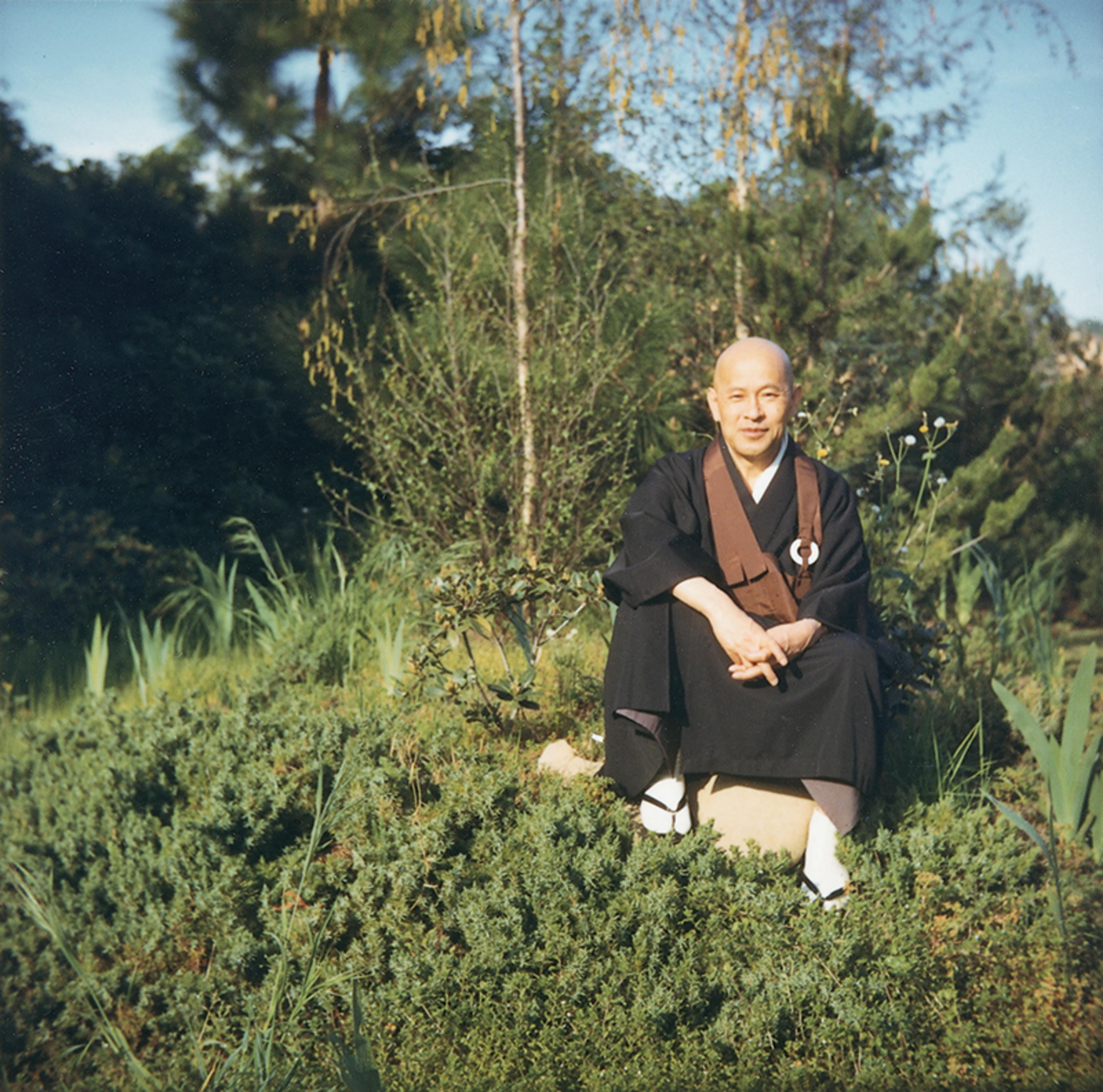 Photo of a person in traditional attire sitting on grass surrounded by greenery and trees, in a tranquil outdoor setting.