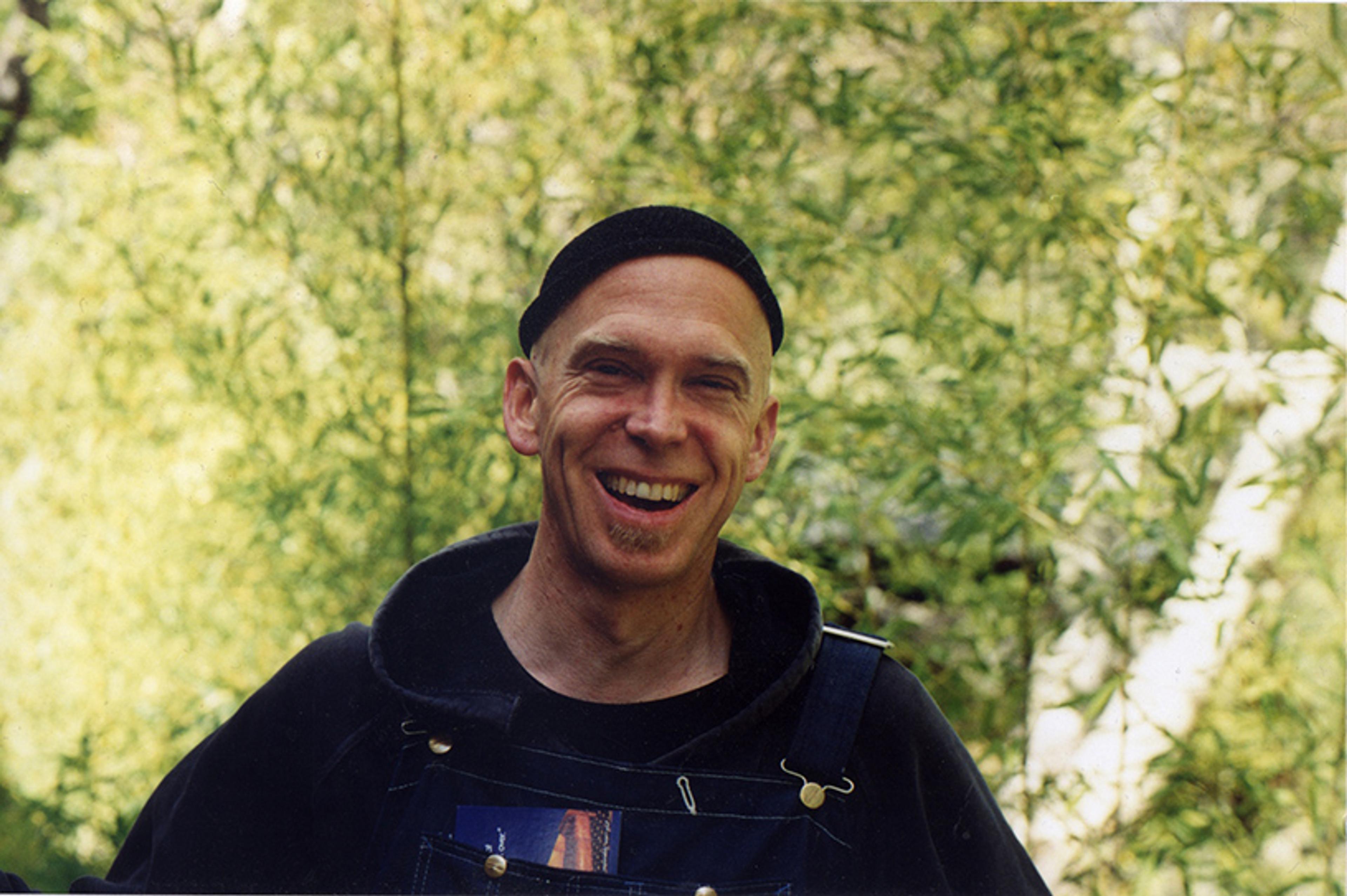 Photo of a smiling man in overalls and a hat standing outdoors with blurred greenery in the background.