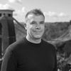 A black and white image of Joe, smiling at the camera. He is outside, with a bridge in the far background, and is wearing a dark-colored sweater.
