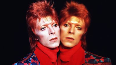 Headshot of David Bowie leaning against a reflective surface. In the reflection, his gaze is direct to the camera. 