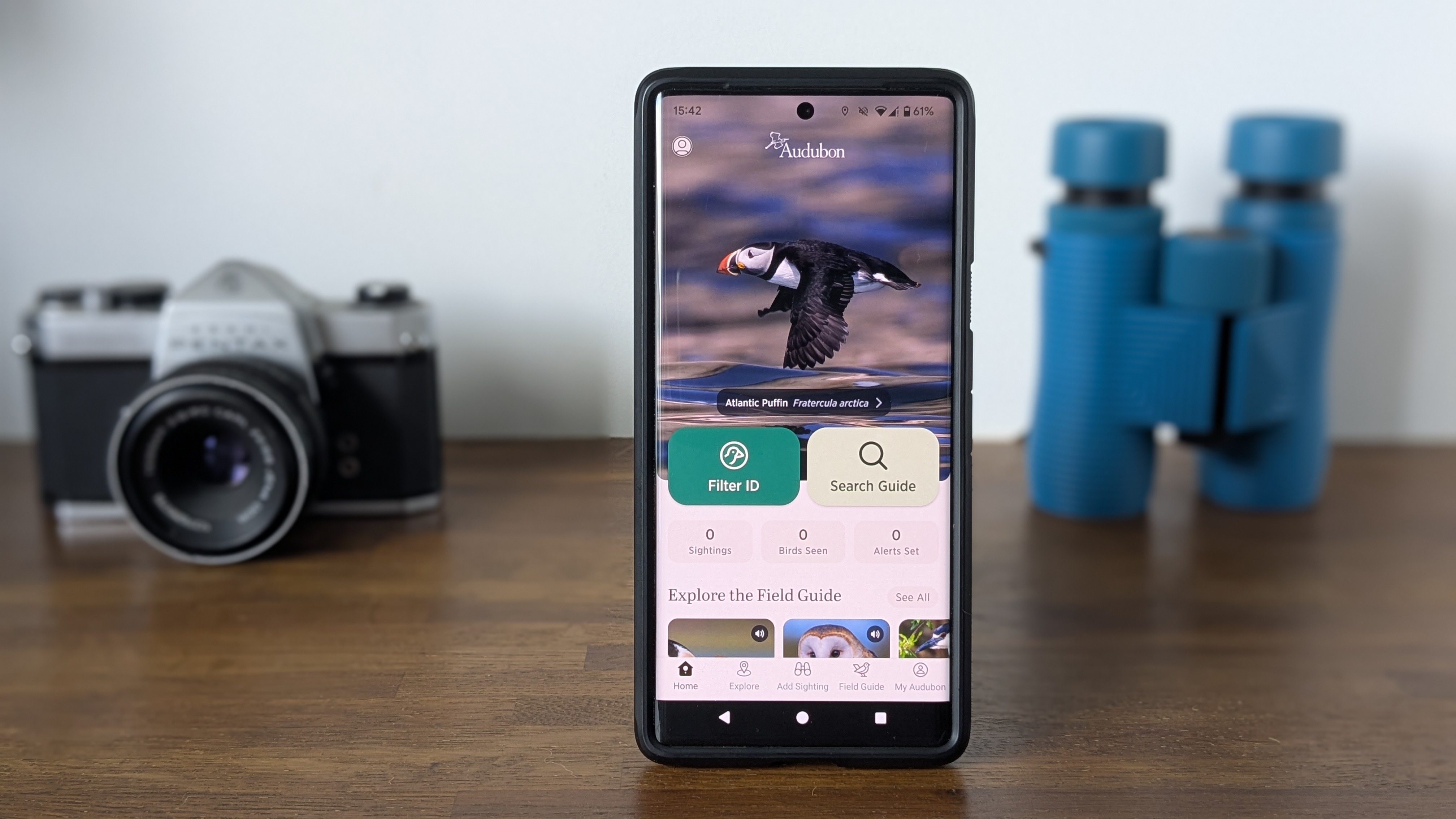 Bird identification app on a wooden table with blue binoculars and a silver SLR camera in the background