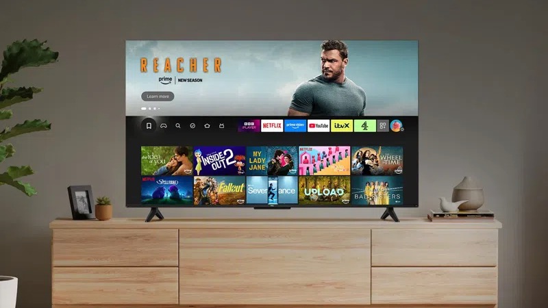 A TCL T6C Fire TV on a wooden media unit. On screen is the Fire TV user interface menu screen.