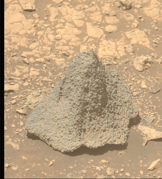 Conical rock on brownish, sandy terrain with a wavy, protruding bottom. It is covered in many small spherical nodules.