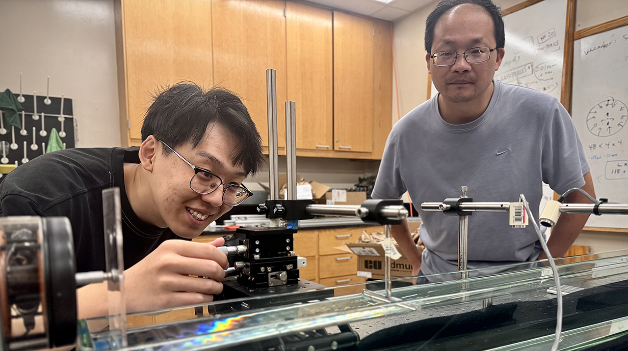 A young man looks through a camera mounted in a track on a laboratory as another man watches.