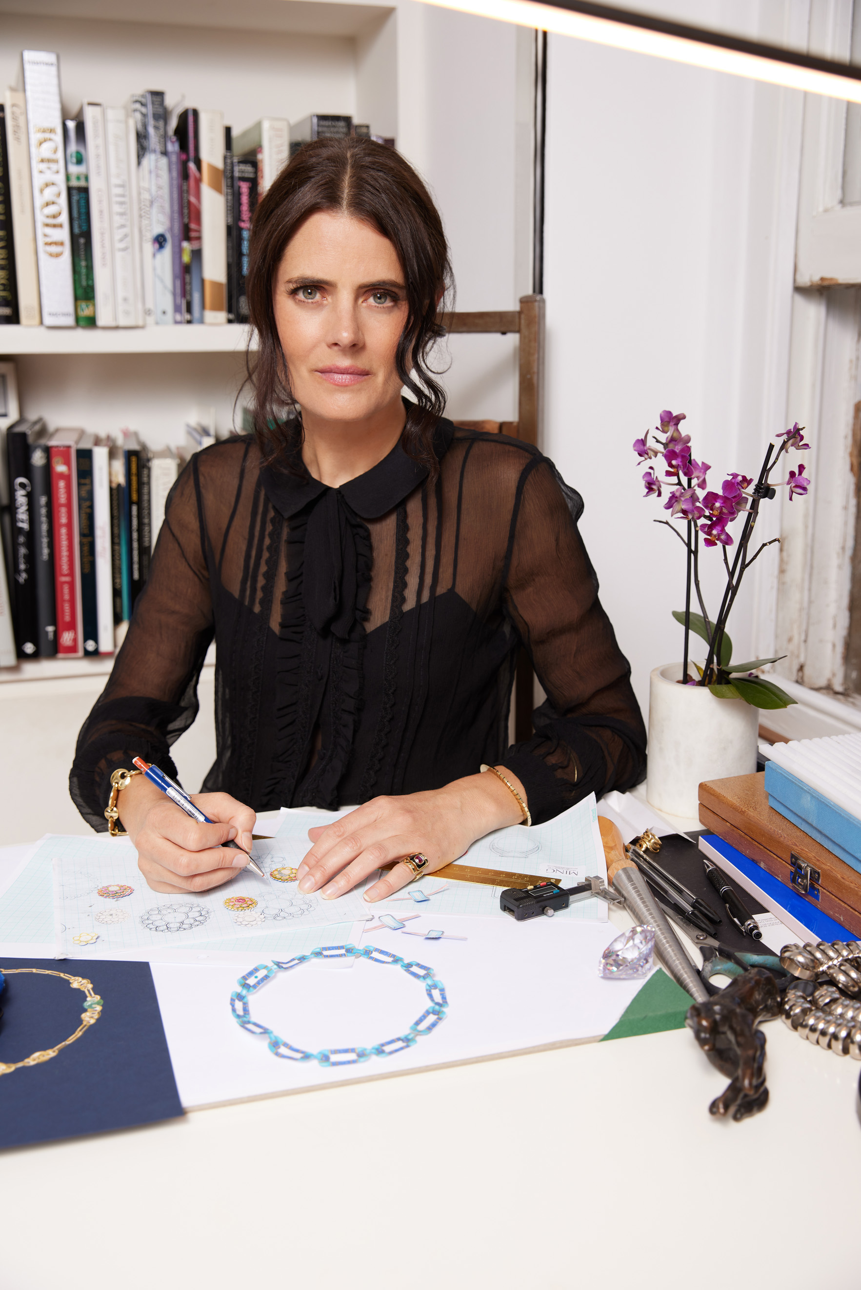 A woman sits at a desk, sketching jewelry designs.