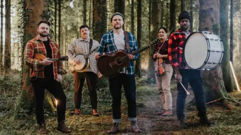 Rend Collective 5 members of Rend Collective standing in a forest. From the left, Wil Pearce has short dark hair and a beard and is holding a ukulele. Next to him, Stephen Mitchell has short, curly black hair and a moustache, and holds a banjo. In the middle, Chris Llewellyn has a beard and a green beanie, and holds a guitar. Next to the right is Jonathan Chu, he has long dark hair and is holding a violin. Next is Daniel Jones wearing a black beanie and holding a large drum strapped around his neck