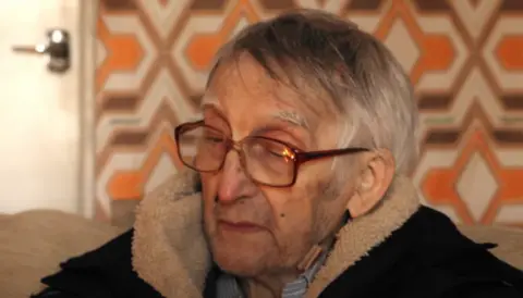 Frances Maguire Owney is sitting down on a sofa, he has short grey hair, red square glasses and a black coat with a cream fleece style around the collar. He is wearing a striped shirt beneath his coat. There is a white door with silver handle behind him, and orange printed wallpaper.